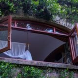 Fereastră larg deschisă într-o cameră luminoasă, cu perdele mișcate de vânt și plante verzi pe pervaz