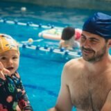 Adult care învață să înoate cu o placă de plutire într-o piscină interioară, ajutat de un instructor