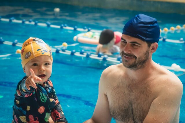 Adult care învață să înoate cu o placă de plutire într-o piscină interioară, ajutat de un instructor