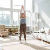 Femeie care stă liniștită lângă o fereastră luminoasă, cu ochii închiși și o mână pe abdomen, respirând lent